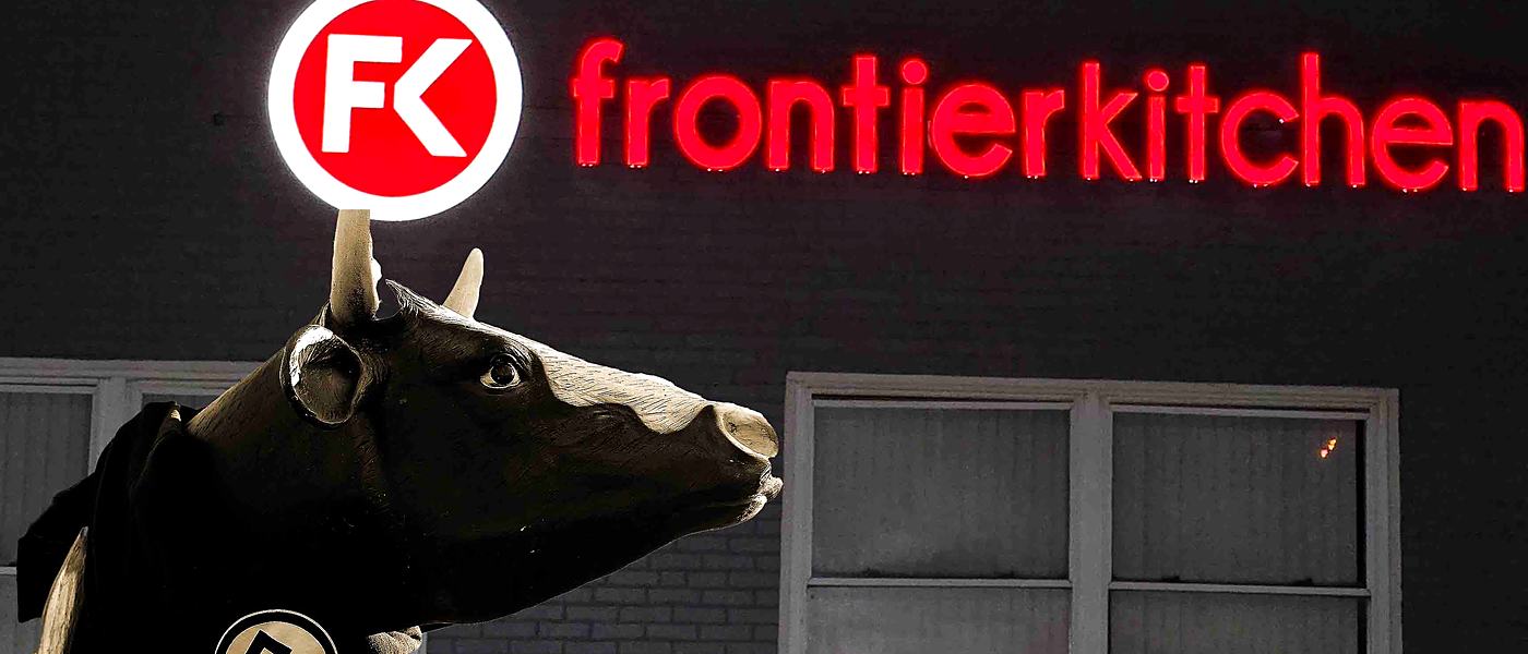 Bessie, a life-size cow statue in a Frontier Kitchen hoodie, standing beneath the lit Frontier Kitchen sign at night.