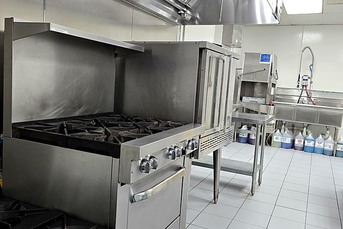 Six-burner commercial gas ranges under an extraction hood, with stainless prep tables alongside.
