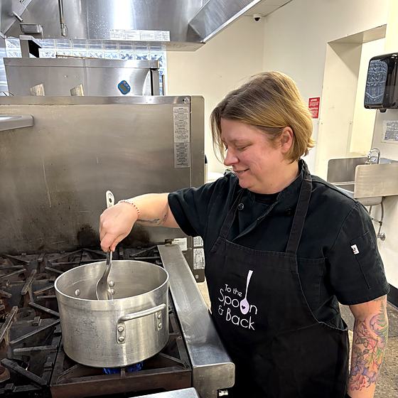 The owner of To The Spoon and Back stirring a pot on a commercial range, wearing a branded apron.