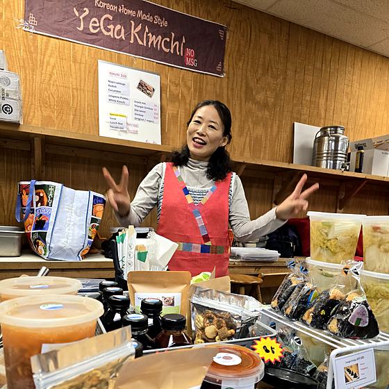 The owner of YeGa Kimchi smiling behind her market stall stocked with jars of kimchi.